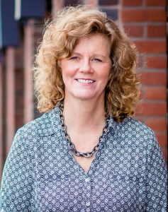 A smiling woman with curly hair wearing a patterned blue shirt and a chain necklace, standing outdoors in front of a brick wall.
